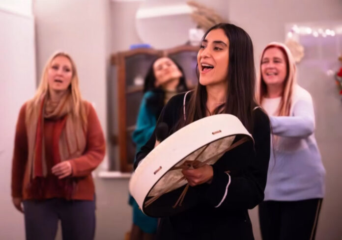 South Asian Woman holding a vegan buffalo drum singing in a circle with 3 people behind her also singing.