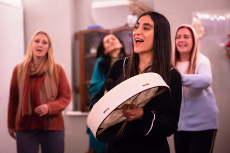 South Asian Woman holding a vegan buffalo drum singing in a circle with 3 people behind her also singing.